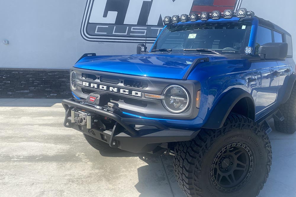 Blue Ford Bronco with aftermarket parts including custom wheels, winch, fender flares, bumper, lights, grill. 